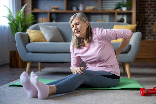 Старшая женщина сидела на коврике для йоги дома и испытывала боль в спине. Нося повседневный костюм тренировки, она иллюстрирует дискомфорт и напряжение с видимым выражением лица. Активный образ жизни.