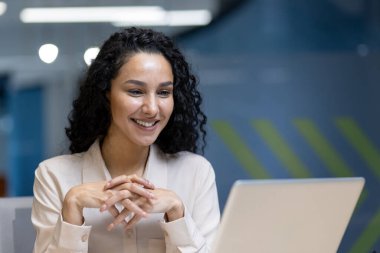 İçerde dizüstü bilgisayarla çalışırken gülümseyen kendinden emin genç bir kadın. Çevrimiçi iletişimle ilgileniyor, kurumsal bir ortamda profesyonel ve olumlu tavırlar sergiliyor..