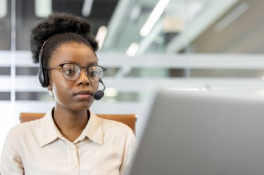 Kulaklıklı kadın parlak ofiste dizüstü bilgisayar kullanıyor. Profesyonelce görevlere yoğunlaşmak, uzaktan kumandayı sergilemek. Modern iletişim ve teknoloji odaklı çalışma ortamı.