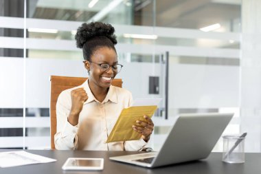 Gülümseyen iş kadını önemli bir belgeyi kutluyor. Masada oturmak, zarfı açmak, sevinç ve memnuniyeti ifade etmek. Sahne dizüstü bilgisayar, akıllı telefon, ofis ayarları, başarı kavramı içerir.