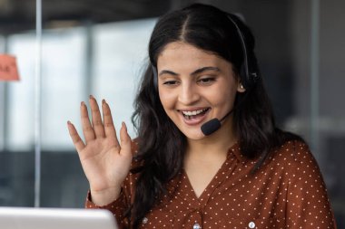 Sanal bir buluşma sırasında kulaklık takan, gülümseyen ve dalgalar sallayan kendine güvenen bir kadın. Takım çalışmasını, iletişimi, uzaktan çalışmayı temsil eder. Profesyonel ortamdaki işbirliği ve bağlantıyı yakalar.