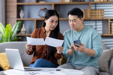 Kanepede oturan Asyalı çift akıllı telefon ve dizüstü bilgisayarla belgeleri inceliyor, finans yönetimini tartışıyorlar. Elinde telefon tutan adam, elinde kağıtlar tutan kadın, rahat oturma odasında takım çalışması..