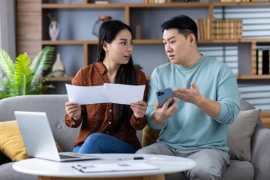 Asyalı çift oturma odasında mali belgeleri telefonla inceliyor. İfadeler şaşkınlık ve endişe gösteriyor. Dizüstü bilgisayarı ve günlük konuşma atmosferiyle modern ev ortamları.