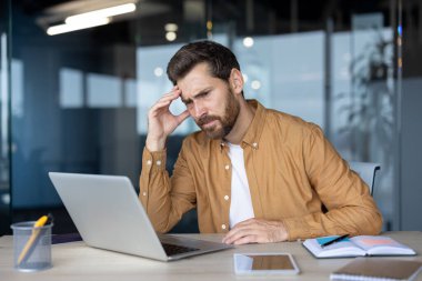 Modern ofis ortamında dizüstü bilgisayarla çalışan stresli bir iş adamı. İnsan zor bir sorun üzerinde düşünüyor gibi görünüyor. Masadaki defter ve telefon teknolojiyi ve iş baskısını vurguluyor.