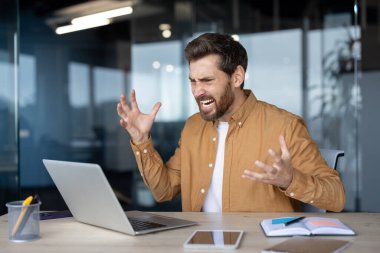 Modern işyerindeki stresli işadamı hayal kırıklığıyla tepki veriyor. Masasında oturan adam açık dizüstü bilgisayar, defter, telefon, ofisteki kızgınlığını ya da memnuniyetsizliğini ifade ediyor..