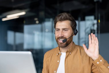 Modern ofiste kendine güveni tam bir adam, kulaklık takıyor, iletişim kuruyor, el sallıyor. Uzaktan çalışmayı, profesyonel etkileşimi, çevrimiçi toplantıyı, dijital bağlantıyı temsil eder.