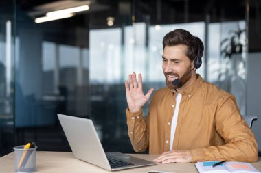 Modern ofis ortamında bilgisayar kulaklığı kullanarak gülümseyen kahverengi gömlekli adam. İş arkadaşlarıyla sanal görüşme yapmak, yenilikçi iletişim teknolojisini göstermek..