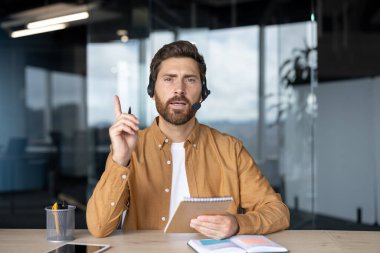 Kulaklıklı bir işadamı sanal toplantı sırasında notlar alıyor, iletişim ve ofis ortamındaki liderliği vurguluyor. Profesyonel sunumu vurgulayarak bir noktaya değinen kişi.