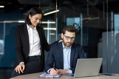 Resmi ofis kılığındaki meslektaşlar masa başında takım çalışması yaparlar. Gülümseyen kadın, düşünceli bir erkekle dizüstü bilgisayar kullanarak iletişim ve iş birliğini vurguluyor..