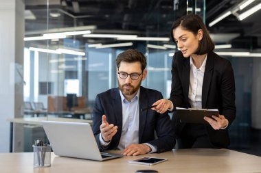 Resmi giyinmiş meslektaşlar iş toplantısındalar. Kadın dizüstü bilgisayarı gösteriyor, erkek çözümü düşünüyor. Bilgisayarlı ofis masası, akıllı telefon ve işbirliğini geliştiren belgeler.