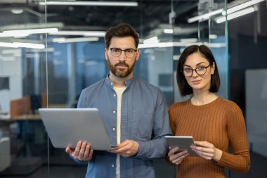 Modern ofis ortamındaki kendine güvenen iş adamları dizüstü bilgisayar ve tablet taşıyorlar. İşbirliği ve teknoloji entegrasyonunu destekleyen dijital çalışma alanına odaklanmış takım çalışması.