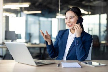 Asyalı iş kadını telefonda çalışırken birden fazla dizüstü bilgisayar kullanıyor ve not alıyor, güven, üretkenlik, teknoloji, iletişim ve profesyonel çalışma ortamını gösteriyor.