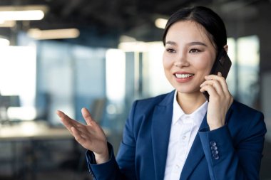 Mavi takım elbiseli Asyalı iş kadını akıllı telefondan konuşuyor. Ofiste işi tartışıyor. Kendine güvenen ifade profesyonelliği, kariyer hırsını, etkili iş iletişimini öneriyor.