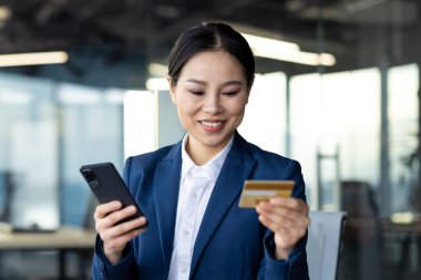 Asyalı iş kadını internet bankacılığı ve ödemeler için akıllı telefon ve kredi kartı kullanıyor. Gülümseyen profesyonel, güven ve modern finans teknolojisini sergiliyor.