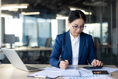 Asyalı iş kadını ofis ortamındaki verileri titizlikle hesaplıyor. Belgeleri analiz ediyor, odaklandığını gösteriyor. Laptop ve hesap makinesiyle donatılmış, modern işyerindeki verimliliği gösteriyor.