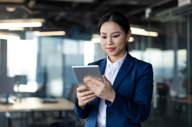 Ofisteki Asyalı iş kadını elinde tablet, takım elbise giymiş, işine odaklanmış. Teknolojiyi, profesyonelliği ve kurumsal çevreye bağlılığı benimsiyor..