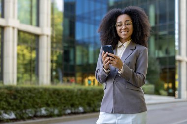 Gri ceketli, güler yüzlü, modern ofis binasının yanında telefonu elinde tutan, kendine güvenen Afro-Amerikan iş kadını. Başarı kavramı, profesyonellik ve dijital iletişim.