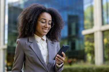Açık havada akıllı telefon kullanan resmi takım elbiseli Afro-Amerikan iş kadını. Gülümsüyor, kendinden emin ve profesyonel görünüyor, şehir ortamında duruyor. Anahtar kelimeler: İş kadını, telefon..