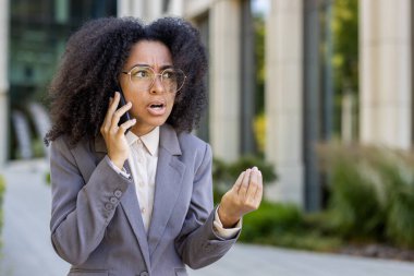 Profesyonel Afro-Amerikan iş kadını gri ceketli gergin bir telefon görüşmesi yapıyor. Onun anlamlı el hareketleri ve endişeli ifadesi hayal kırıklığını, aciliyeti ve iş stresini ifade eder..