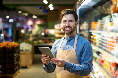 Market çalışanı süpermarket koridorunda önlük giyiyor. Gülümseyen adam, envanteri yönetmek ve müşteri hizmetlerini geliştirmek için teknolojiyi kullanıyor..
