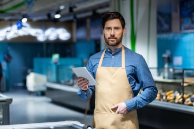 Modern süpermarkette önlüğü ve tableti olan bir tezgahtar müşterilere alışveriş ihtiyaçları konusunda yardım etmeye hazır. Canlı perakende ortamında kendinden emin ve etkili..