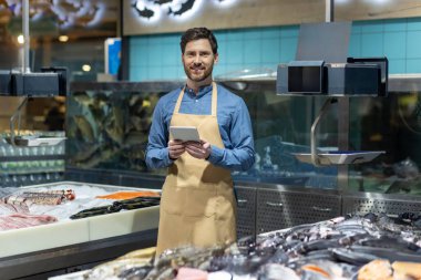 Deniz ürünleri reyonunda önlüklü erkek balıkçı. Elinde tabletle gülümsemek, envanter yönetimi, tazelik ve market ortamında teknoloji kullanımını sergilemek.