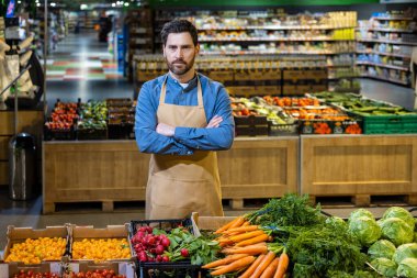 Önlüklü bir market çalışanı kolları bağlı duruyor. Modern süpermarkette havuç ve domates gibi taze ürünlerle çevrili. Işıl ışıl bir iç mekan.