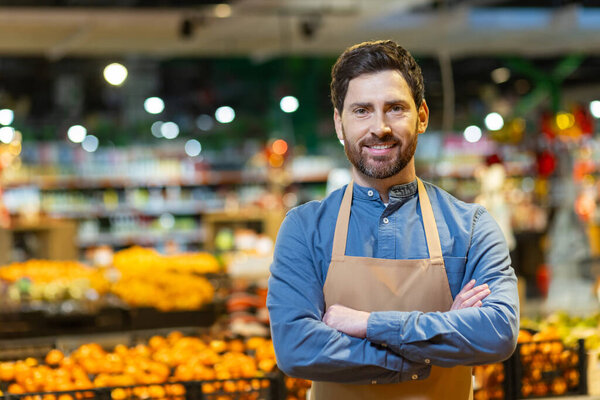 Confident male grocery worker wearing apron stands with crossed arms in front of fresh produce section at supermarket. Smiling expression conveys professionalism, hospitality, dedication.