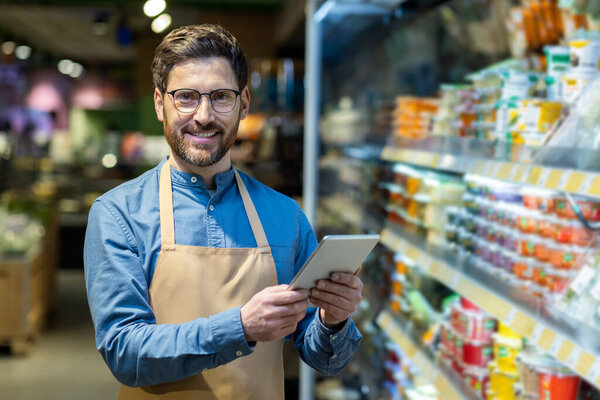 Grocery store worker in apron smiling while using digital tablet for inventory management. Enhancing customer service with technology in retail setting. Efficient and friendly service