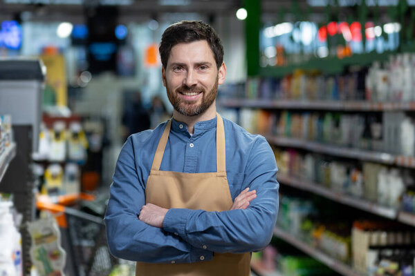 Confident male employee with apron stands with crossed arms in grocery store aisle, exuding professionalism and customer service expertise, surrounded by variety of products in retail setting.