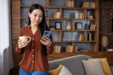 Asyalı kadın akıllı telefon ve kahve tutuyor, kitaplığı olan rahat bir kanepenin yanında duruyor. Evde rahat bir ortam, telefonda mesaj okumak, evde boş vakitlerin tadını çıkarmak..