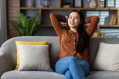 Asyalı kadın modern oturma odasında koltukta dinleniyor, memnuniyeti ve huzuru kucaklıyor. Rastgele bir şekilde evinde huzurlu anlar yaşa..