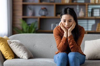 Asyalı kadın gri koltukta oturur düşünceli ve yalnız hisseder. Modern dekorlu, yastıklı rahat bir oturma odası. İletken duygular arasında tefekkür, yalnızlık, yansıma, farkındalık vardır..