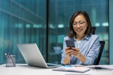 Asyalı iş kadını masa başında akıllı telefon kullanıyor, dizüstü bilgisayarda çalışıyor, modern ofiste gülümsüyor. Dijital üretkenliği, iletişimi, teknolojiyi ön plana çıkarıyor. İş ve iş temaları için ideal.