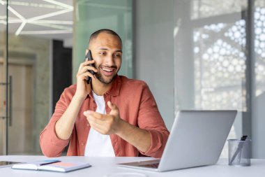 Kendine güvenen bir iş adamı, ofis alanındaki dizüstü bilgisayarını kullanarak birden fazla görev ve iletişim becerilerini aktarıyor. Günlük kıyafet rahat ama verimli bir çevreyi yansıtır..
