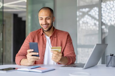 Ofis masasında online alışveriş yaparken elinde akıllı telefon ve kredi kartı tutan gülümseyen adam. Telefonla dijital alışverişe girişmek, konfor ve modern teknolojiyi sergilemek.