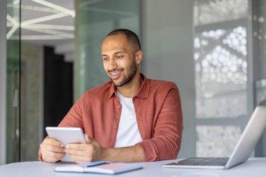 Modern ofis ortamında dijital tablet kullanan gülümseyen adam. Dijital görevleri tablet ve dizüstü bilgisayarla gerçekleştirir. Günlük giysiler rahat ama profesyonel bir atmosferi işaret ediyor..