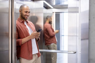 Asansörde akıllı telefon kullanan, ayna duvarına yaslanan, çaba harcamadan gülümseyen, günlük kıyafetler giyen bir adam. Sahne, bağlantı, iletişim, modern teknoloji, günlük rutin tasvir eder.