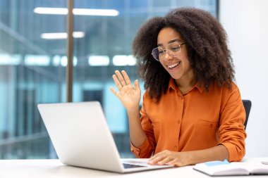 Smiling woman in orange shirt waves during online meeting, engaging in communication through laptop. Bright office setting conveys positive work-from-home environment and internet connectivity. clipart