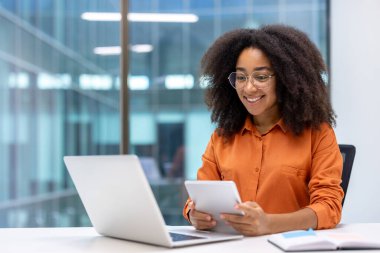 Turuncu tişörtlü gülümseyen kadın masadaki dijital tablette dizüstü bilgisayarla çalışıyor. Teknoloji ve dijital bağlantı odaklı modern ofis ortamında profesyonel ortam.