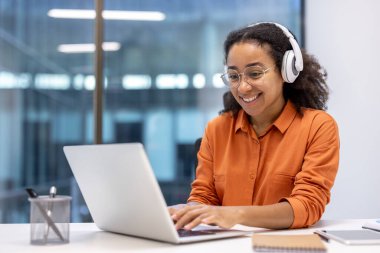 Kulaklık takan kadın programcı, dizüstü bilgisayarda yeni yazılım kodluyor. Modern ofiste çalışırken odaklanmış ve mutlu. Teknoloji, iş kavramları ve ilham için ideal..
