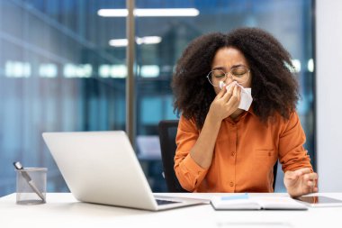 Ofisteki kadın, masa başı mendil kullanarak hapşırıyor. Soğuk algınlığı belirtilerini ve sağlık bilincini gösteriyor. Hastalık sırasında çalışma kavramı ve profesyonel ortamlarda kendi kendine bakım ihtiyacı.
