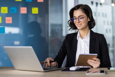 Siyah takım elbiseli kendine güvenen iş kadını proje için dizüstü bilgisayar kullanıyor. Elinde renkli yapışkan notlarla ofiste defter tutarken gülümsüyorsun. Çevrimiçi iş iletişimi.
