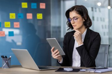 Ofisteki iş kadını, renkli yapışkan notlarla çevrili tablet ve laptopları analiz ediyor. Modern çalışma alanında odaklanmış profesyonel projeler güven ve üretkenlik.