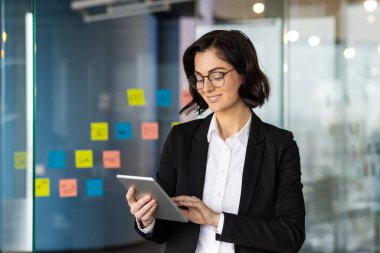 Takım elbiseli kendine güvenen profesyonel kadın modern ofis ortamında dijital tablet kullanıyor. Etrafı cam duvardaki renkli yapışkan notlarla çevrili, proje yönetimi ve verimliliğine odaklanmış..
