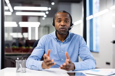 Modern ofiste kulaklık takan olgun bir işadamı iş stratejilerini tartışırken görüntülü konuşma yapıyor. Profesyonel iş ortamında sakin ve odaklanmış tavır