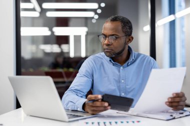 Olgun Afro-Amerikan iş adamı vergi formlarını inceliyor, hesap makinesi tutuyor. Bilgisayarın yanı sıra hesaplamalar için akıllı telefon kullanıyor. Sahne aktarımları odak, analiz, mali yönetim.