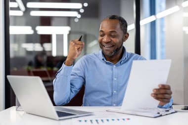Neşeli Afro-Amerikalı olgun iş adamı ofis masasında başarıyı laptop ve belgelerle kutluyor, iş başarısını ve kariyer ilerlemesine olan hevesini yansıtıyor..