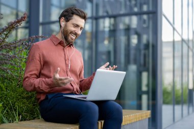 İş adamı dışarıda dijital toplantı için dizüstü bilgisayar kullanıyor. Kulaklıkla günlük kıyafetlerle gülümsüyor, modern bina arka planına sahip bankta oturuyor. İletişimi, teknolojiyi, başarıyı gösterir.