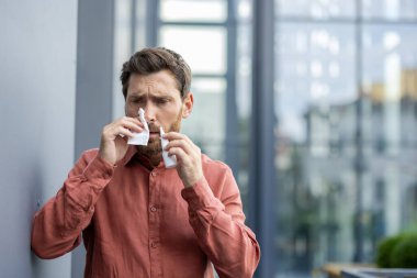 Şehir ortamında dokuları kullanan, günlük gömlekler giyen, endişeli görünen bir adam. Sahne, sağlık ve hijyendeki günlük zorlukları yansıtan soğuk algınlığı veya alerji gibi sağlık sorunlarını betimliyor..